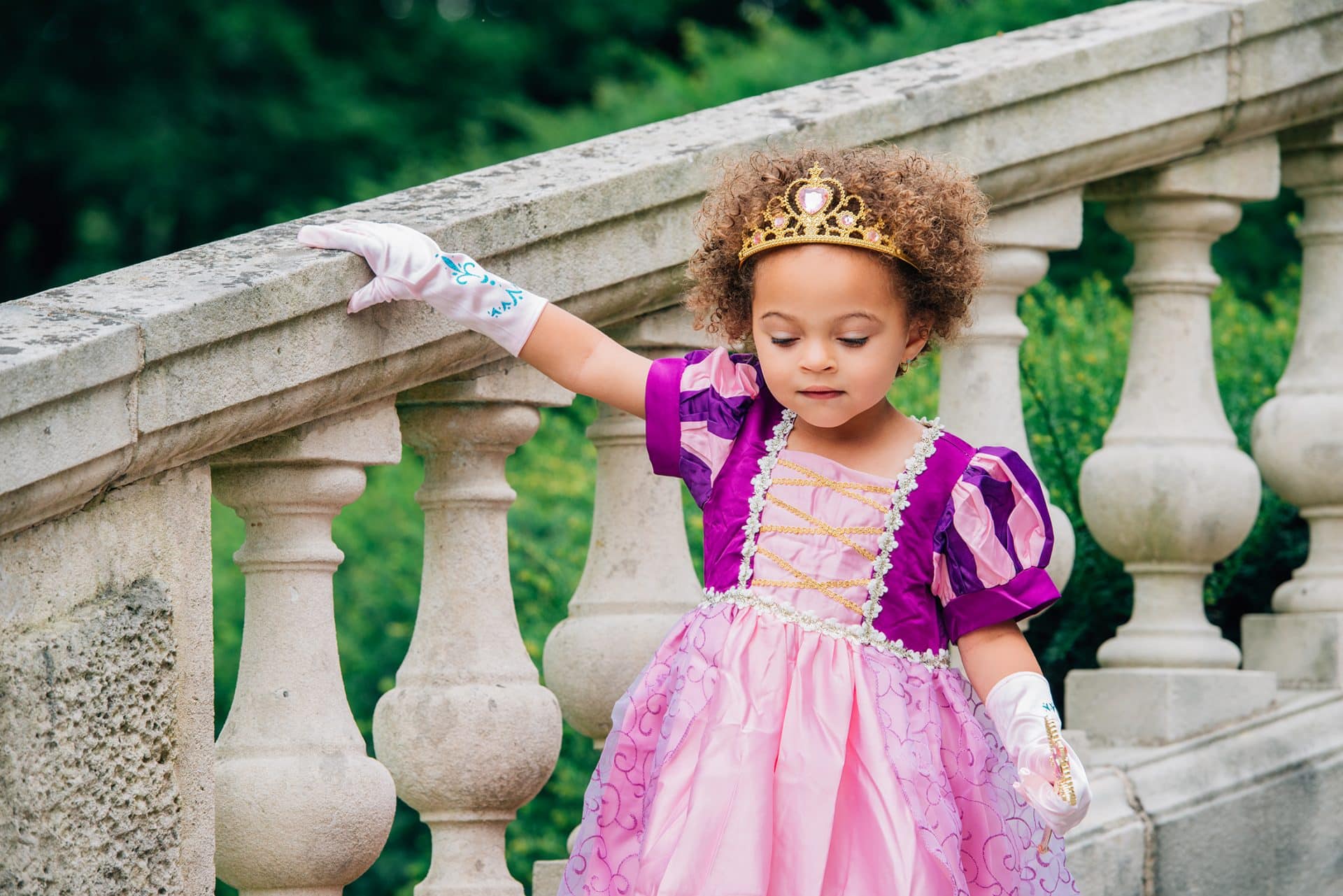 Meisje draagt het rapunzel kostuum van Het Betere Merk met paarse top, roze rok, tiara en witte handschoenen bij een balustrade.