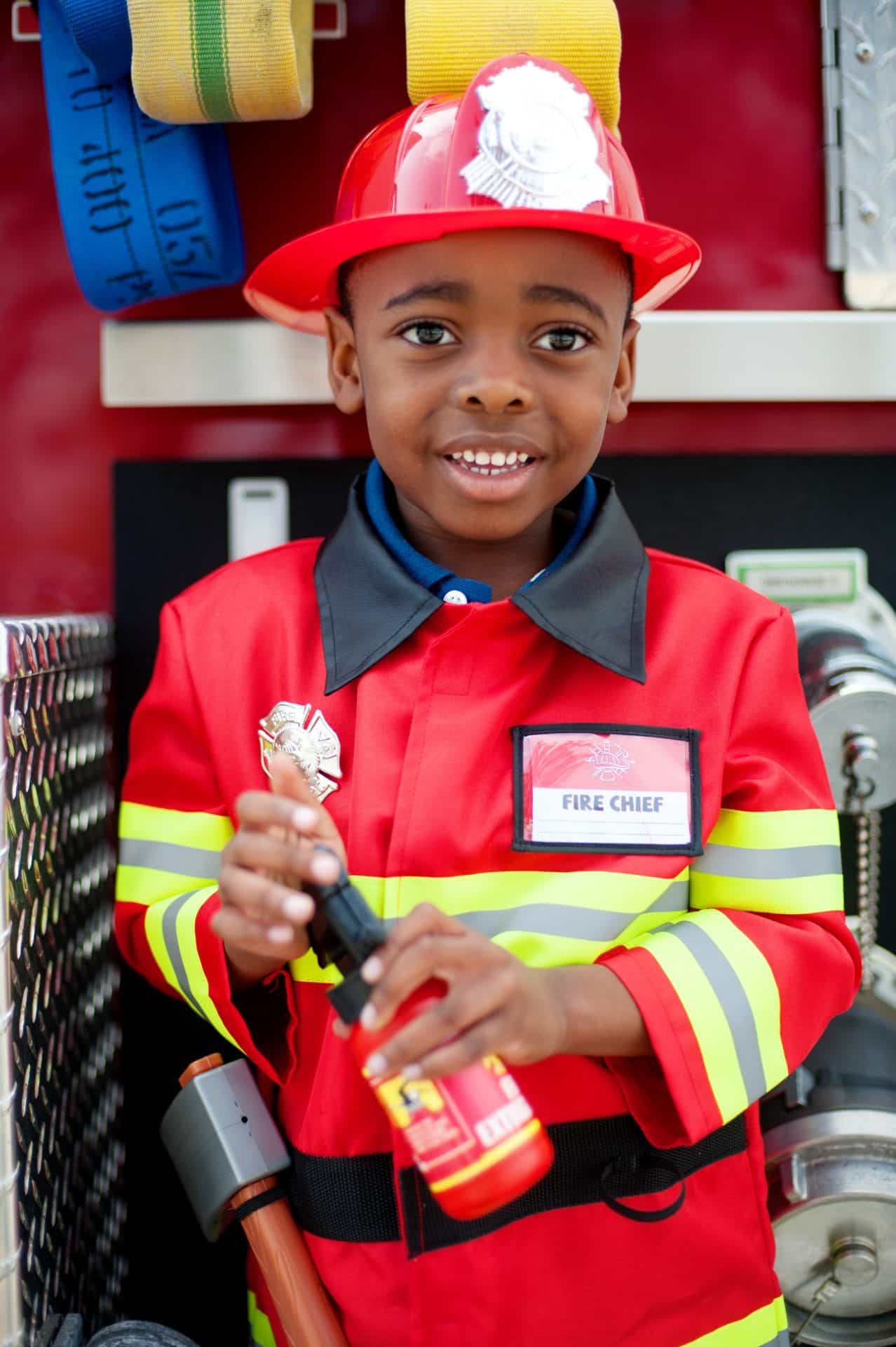 Jongen draagt een rood brandweer kostuum met reflecterende strepen, brandweerhelm en naamplaatje, terwijl hij een blusser vasthoudt.