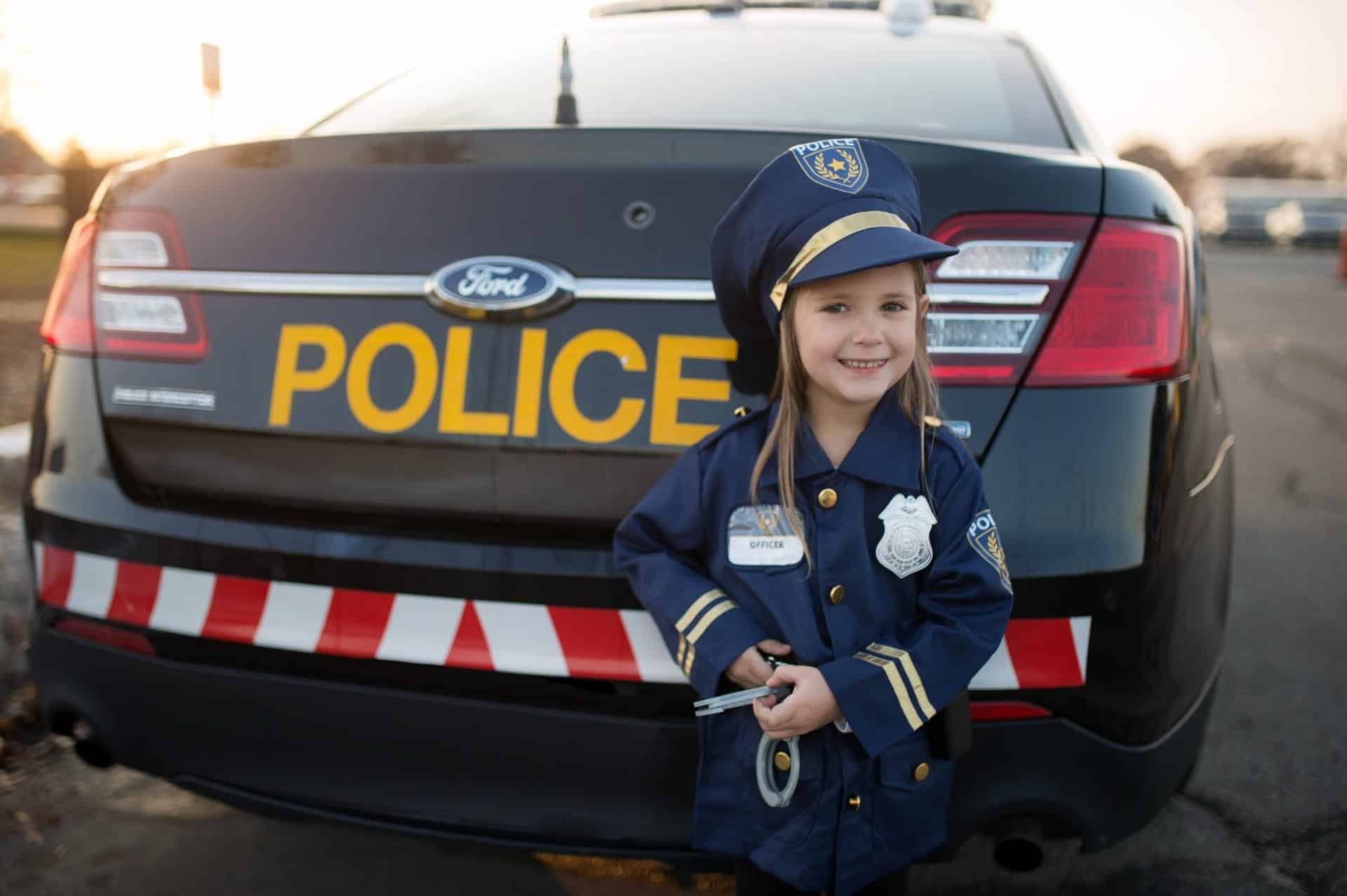 Meisje draagt een donkerblauwe verkleedset politie kind met pet en handboeien voor een politieauto.