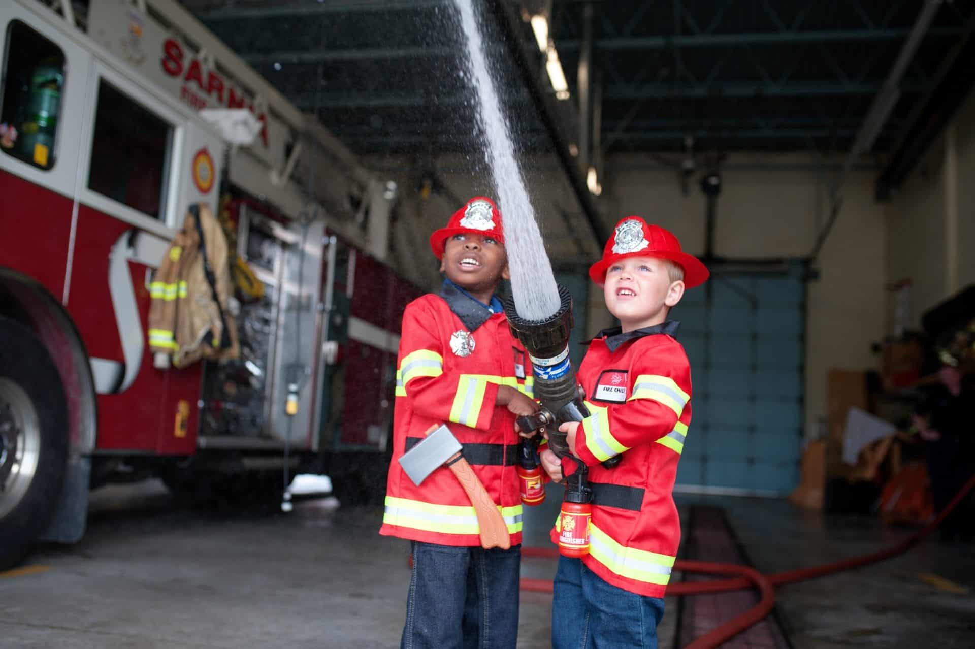 Twee jongens dragen een rood brandweerkostuum met reflecterende strepen en brandweerhelmen terwijl ze samen een slang vasthouden.