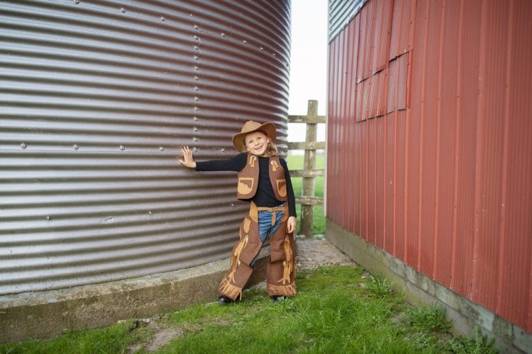 Kind draagt een cowboy outfit met bruin vest, cowboyhoed en beenbeschermers tussen schuren