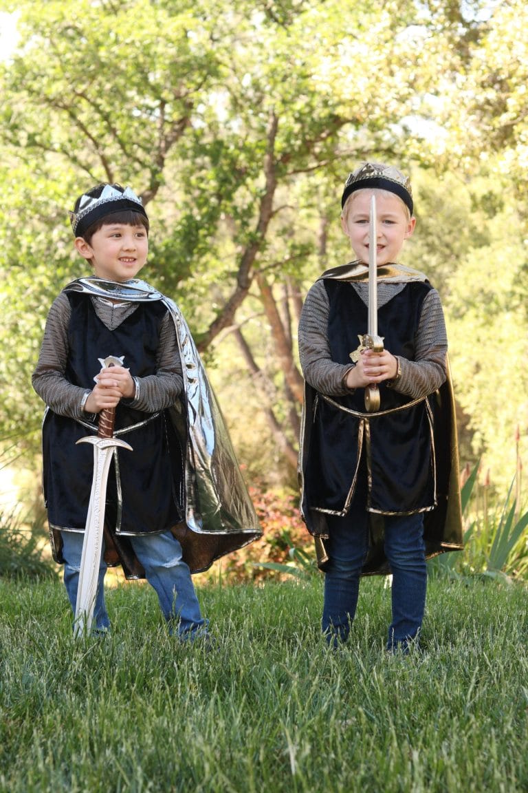 Twee jongens dragen Great Pretenders verkleedkleding als ridders met kronen, capes en zwaarden in een groene tuin.