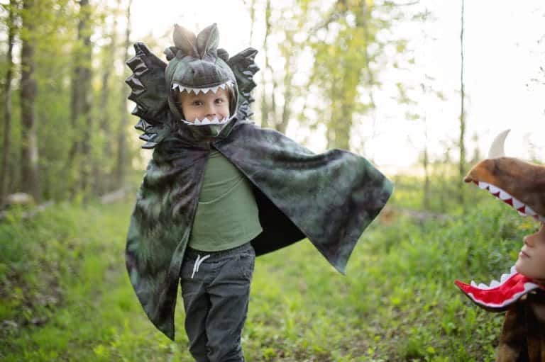 Kind draagt een groen-grijze dino cape met dinosauruskop en witte tanden in het bos, naast een ander kind in kostuum