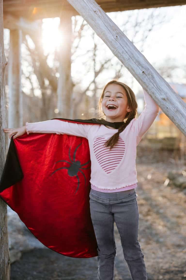 Meisje draagt de spiderman batman cape met rood spinnenembleem buiten aan een houten balk
