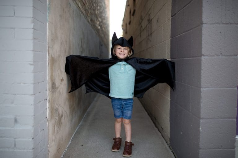 Een kind draagt een zwarte batman cape met capuchon en vleermuisoren, met de armen wijd uitgespreid.