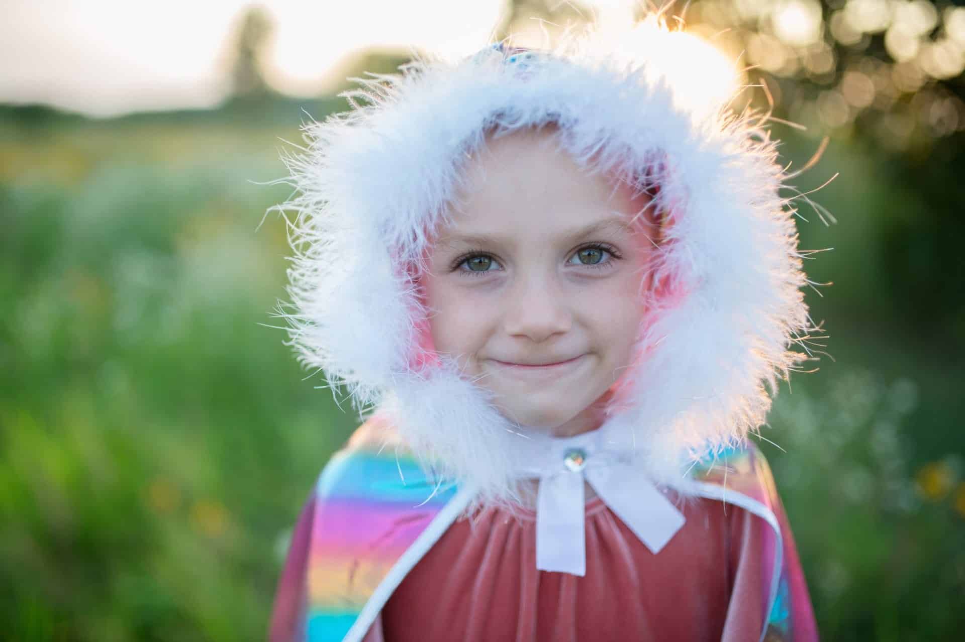 Kind draagt de holografische roze prinsessencape met witte bontkraag en capuchon, buiten in het groen