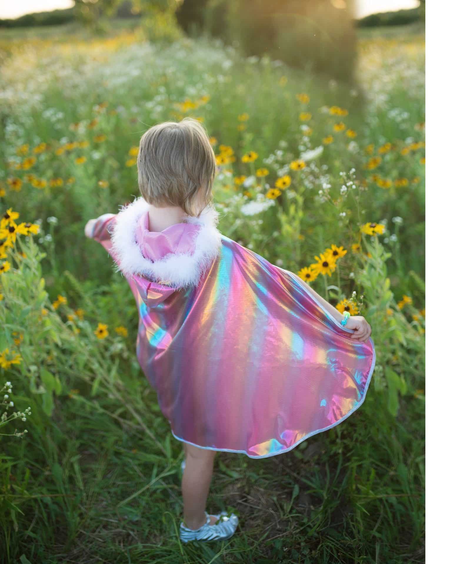 Van achteren spreidt een kind de holografische roze prinsessencape met witte bontkraag in een bloemenweide.