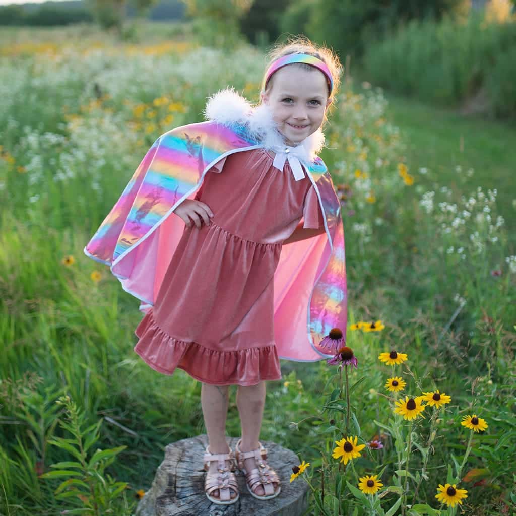 Meisje draagt een roze prinsessencape met holografische regenboogglans over een roze jurk tussen bloemen