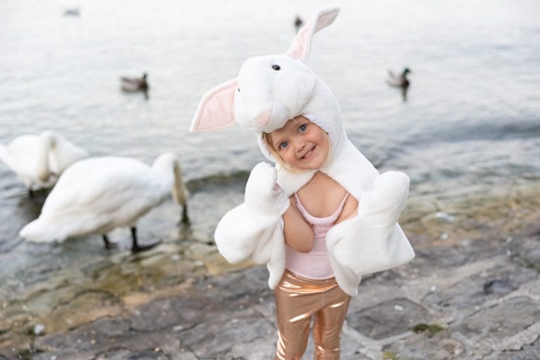Kind draagt een witte konijnen cape met konijnenkop en roze oren bij het water, met zwanen op de achtergrond.