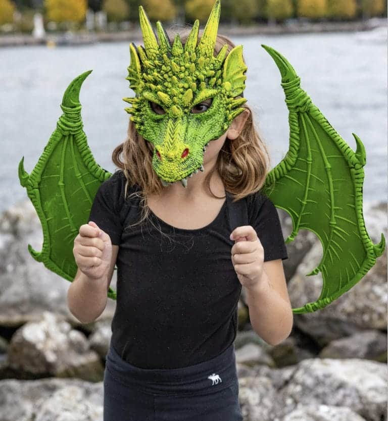Kind draagt een groen draken masker met stekels, hoorns en scherpe tanden; grote groene vleugels zijn zichtbaar.