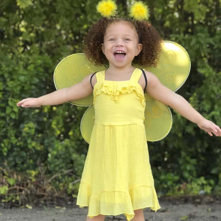 Kind draagt gele Great Pretenders hommel vleugels met bijpassende hoofdband en gele jurk in de tuin