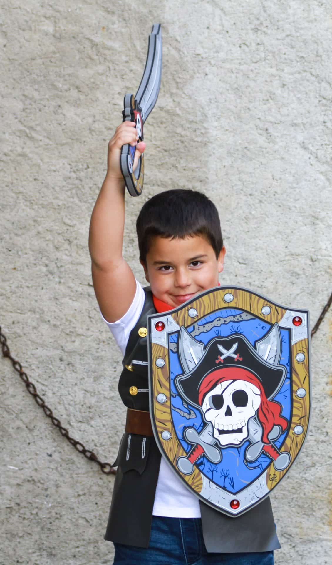 Jongen houdt een Great Pretenders piratenzwaard omhoog en draagt een blauw piratenschild met doodskop.