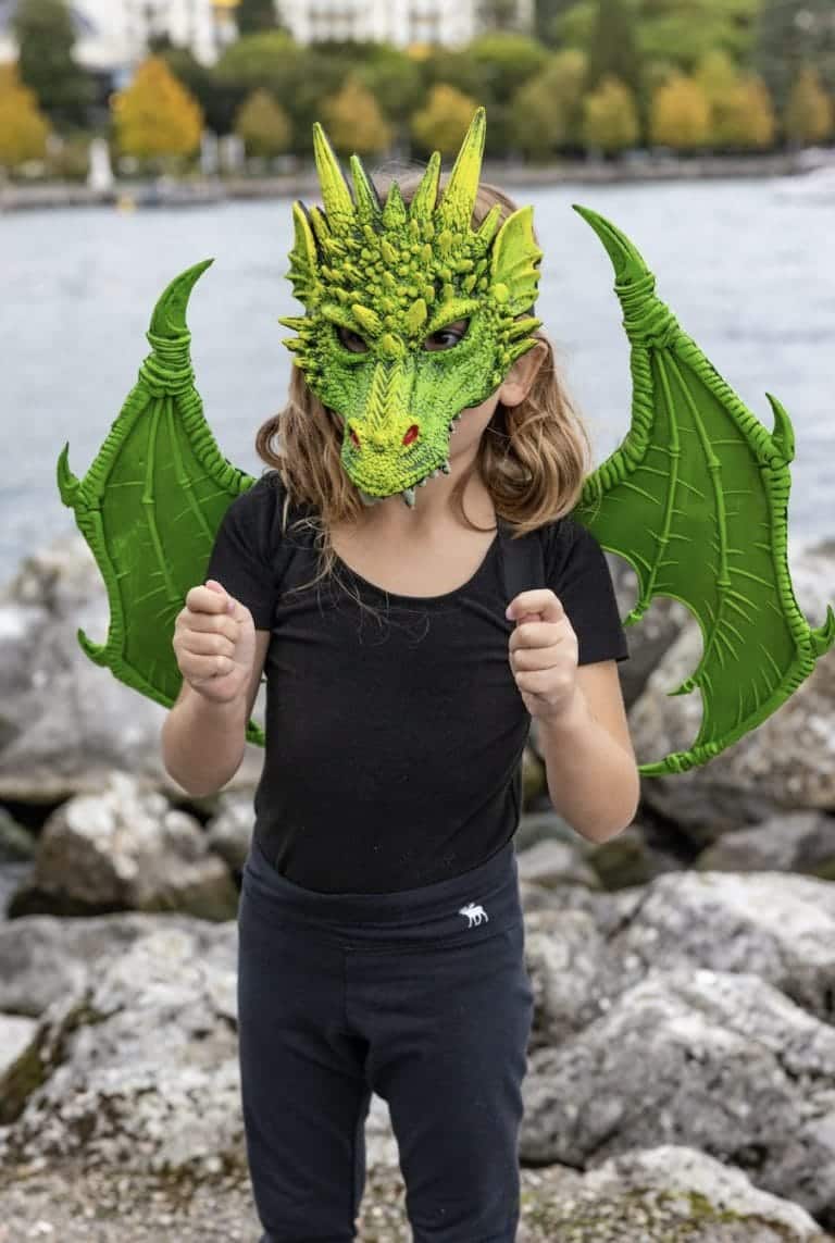 Kind draagt groene draken vleugels met een groen drakenmasker, staand buiten bij het water