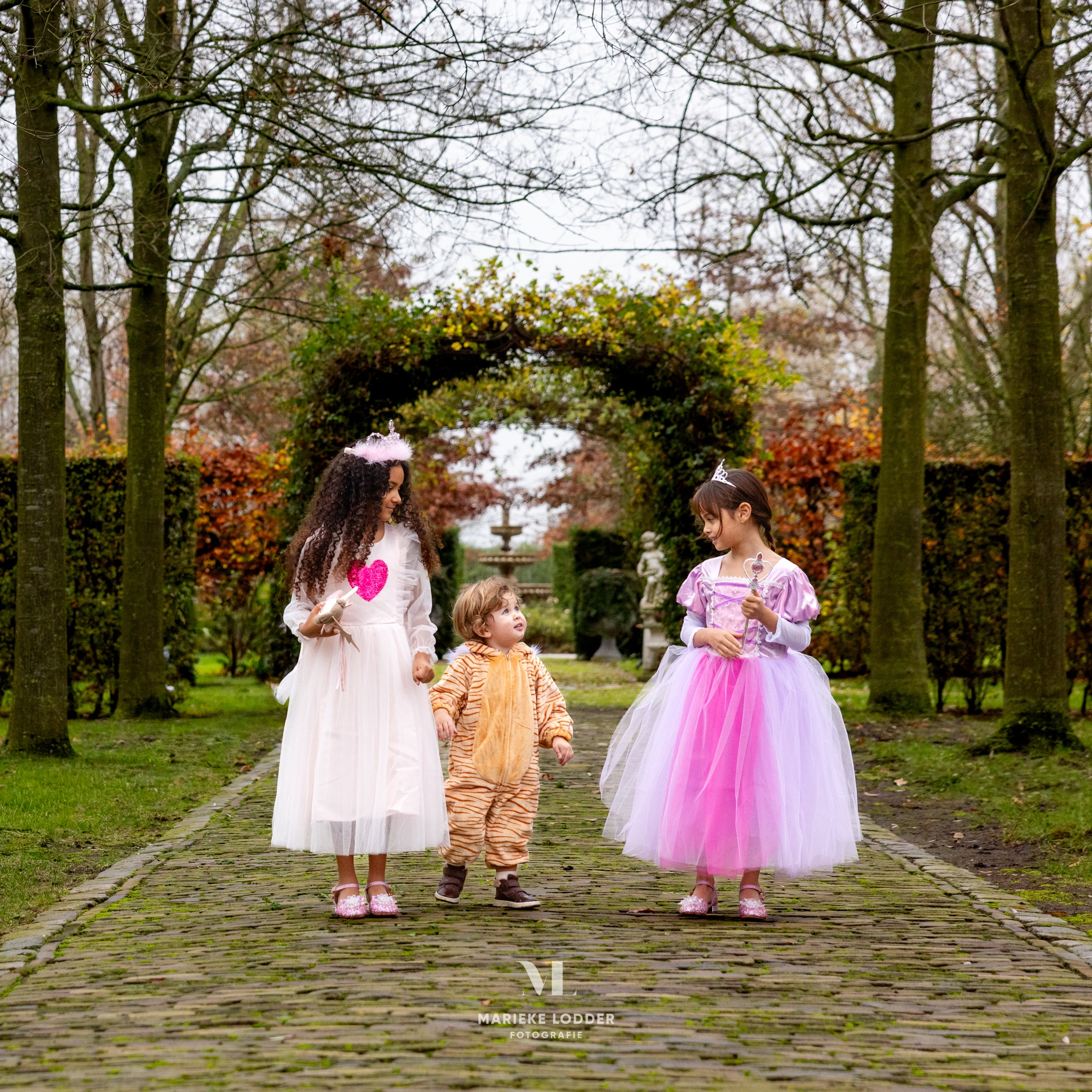 Kind in een tijger onesie met oranje vacht en donkere strepen loopt tussen twee meisjes in prinsessenjurken door een tuin.