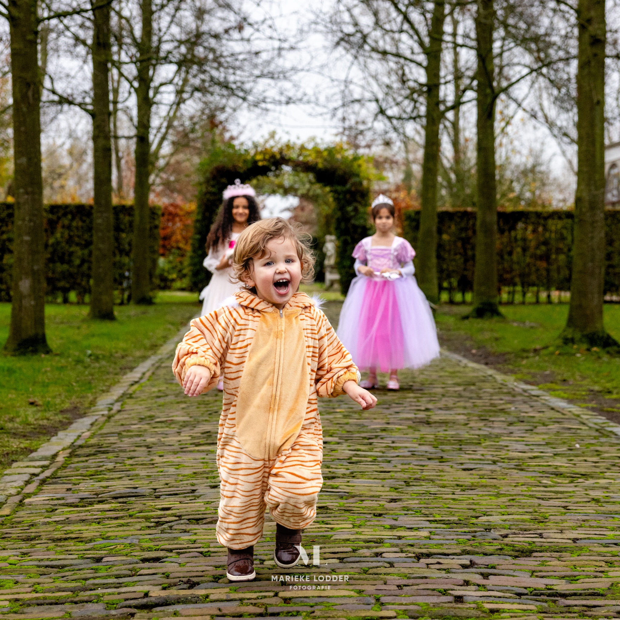 Kind draagt een oranje tijger onesie met donkere strepen en rent over een klinkerpad, met twee prinsessen op de achtergrond