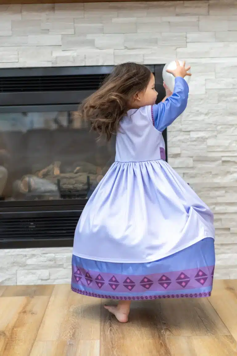 Meisje in de Wens ster prinsessenjurk draait zich om bij een open haard, met een witte bal en paarse zoomversiering.