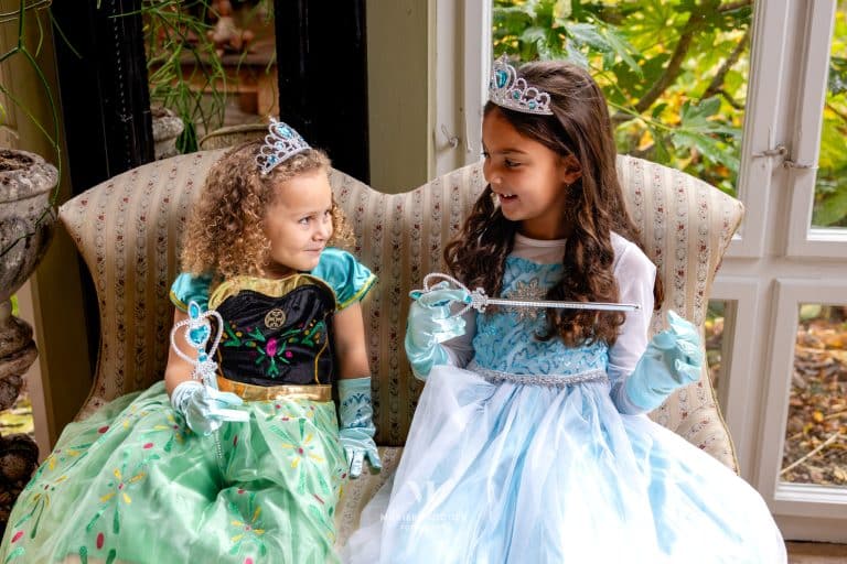 Twee meisjes dragen de Anna- en Elsa-jurk met kroontjes en bijpassende handschoenen, zittend op een bank.