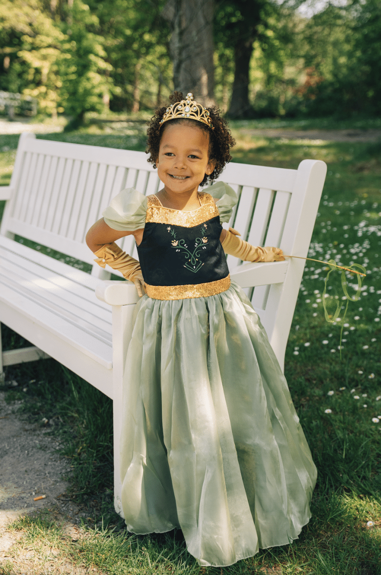 Meisje draagt een groene prinsessenjurk van Anna met gouden details, handschoenen en kroon op een witte bank in het park.