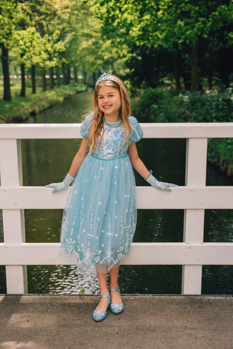 Meisje draagt de blauwe Elsa prinsessenjurk van Het Betere Merk met handschoenen, tiara en bijpassende schoenen op een brug.