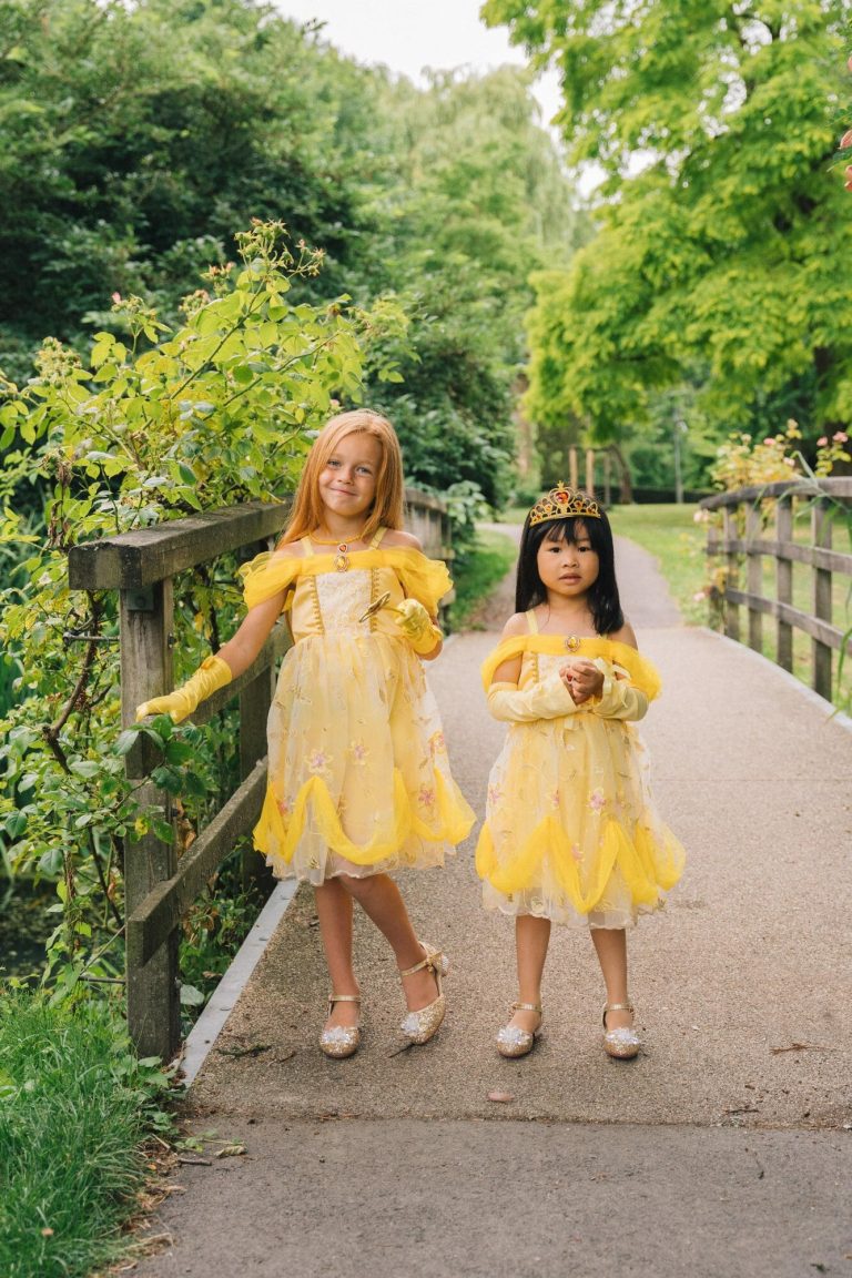 Twee meisjes dragen een gele Belle prinsessenjurk met glanzende schoenen; één meisje draagt een kroon op een groen pad.