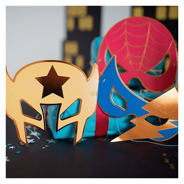 Superhelden maskers in goud, blauw en roze, met sterren, bliksemschicht en spinnenwebmotief op tafel.