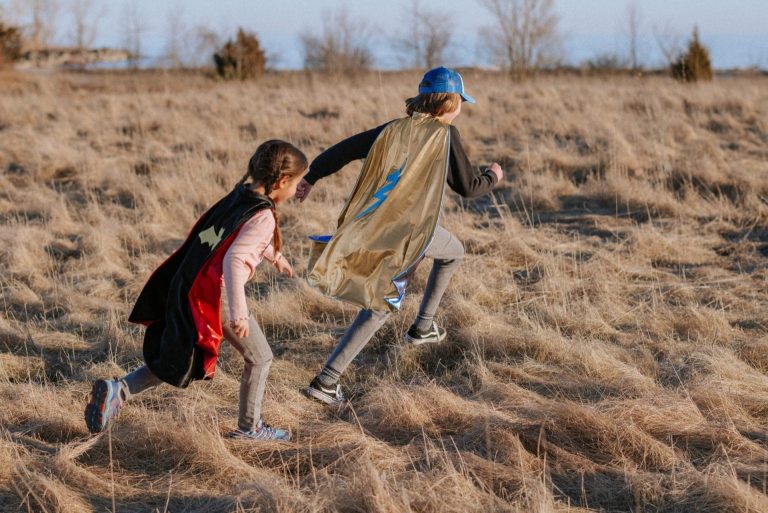 Twee kinderen rennen buiten met een zwarte en gouden superhelden cape, waarvan één met blauwe bliksemschicht