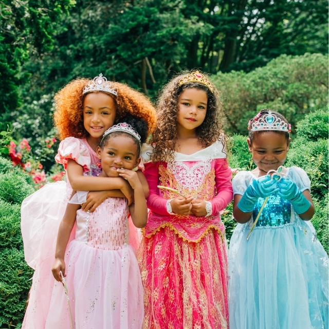 Vier meisjes dragen kleurrijke prinsessenjurken met kroontjes als carnavalskleding meisjes in een groene tuin.
