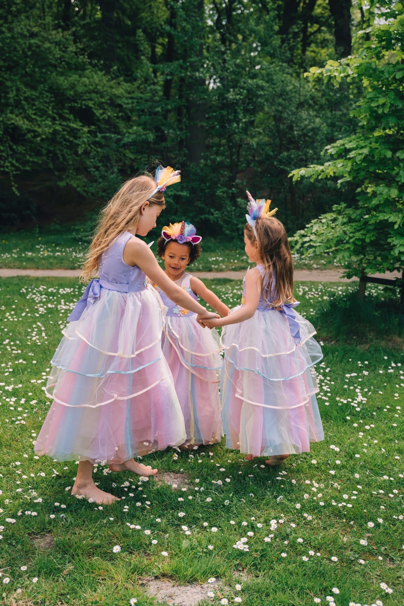 Drie meisjes houden elkaar vast in lila unicorn jurkjes met lange tule rokken en kleurrijke haarbanden op een grasveld.