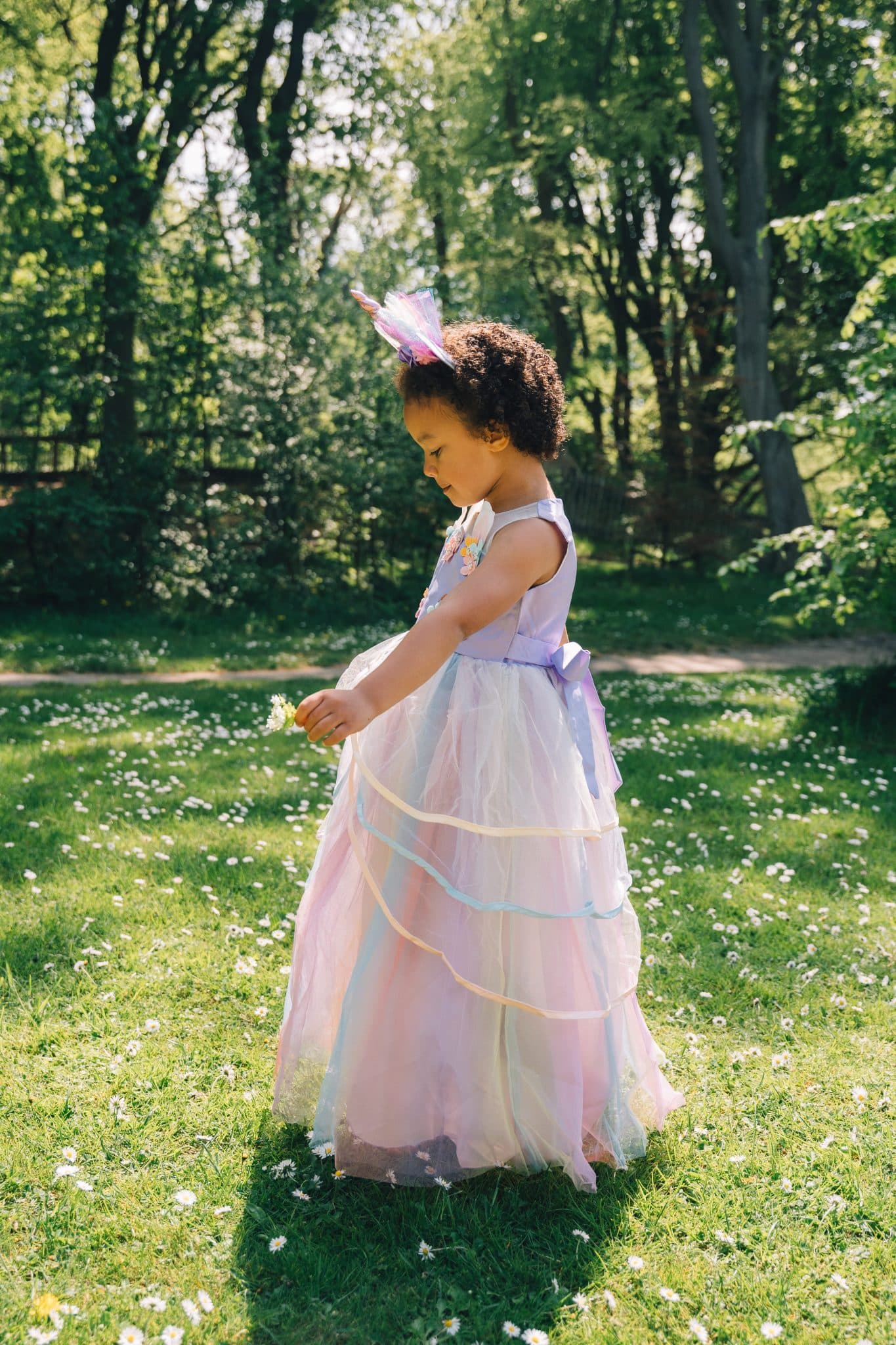 In zijaanzicht draagt een kind de lila unicorn jurk met gelaagde tule rok en haarband op een zonnige bosweide.