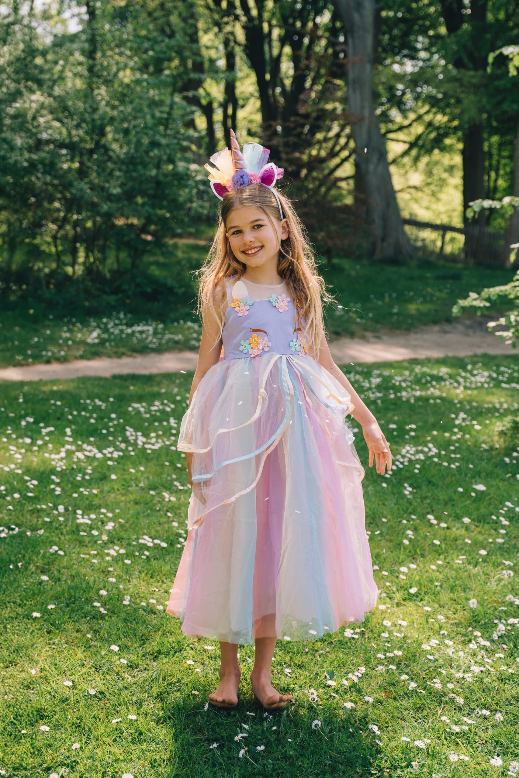Meisje draagt een lila unicorn jurk met kleurrijke haarband en transparante tule rok in een bloemenweide.
