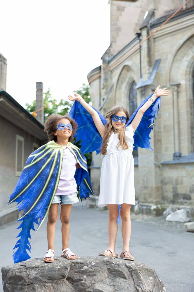 Twee kinderen dragen blauwe draken capes en maskers, met uitgespreide vleugels buiten bij een stenen gebouw.
