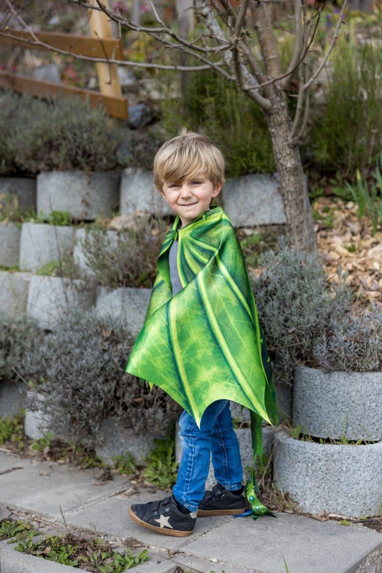 Jongen draagt een groene drakencape met vleugelpatroon en puntige onderkant in een tuin.