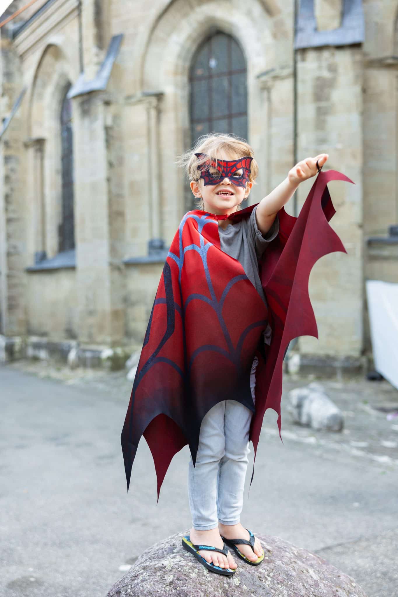 Jongen draagt een rode Spiderman-cape met masker en houdt een vleugel omhoog voor een stenen gebouw