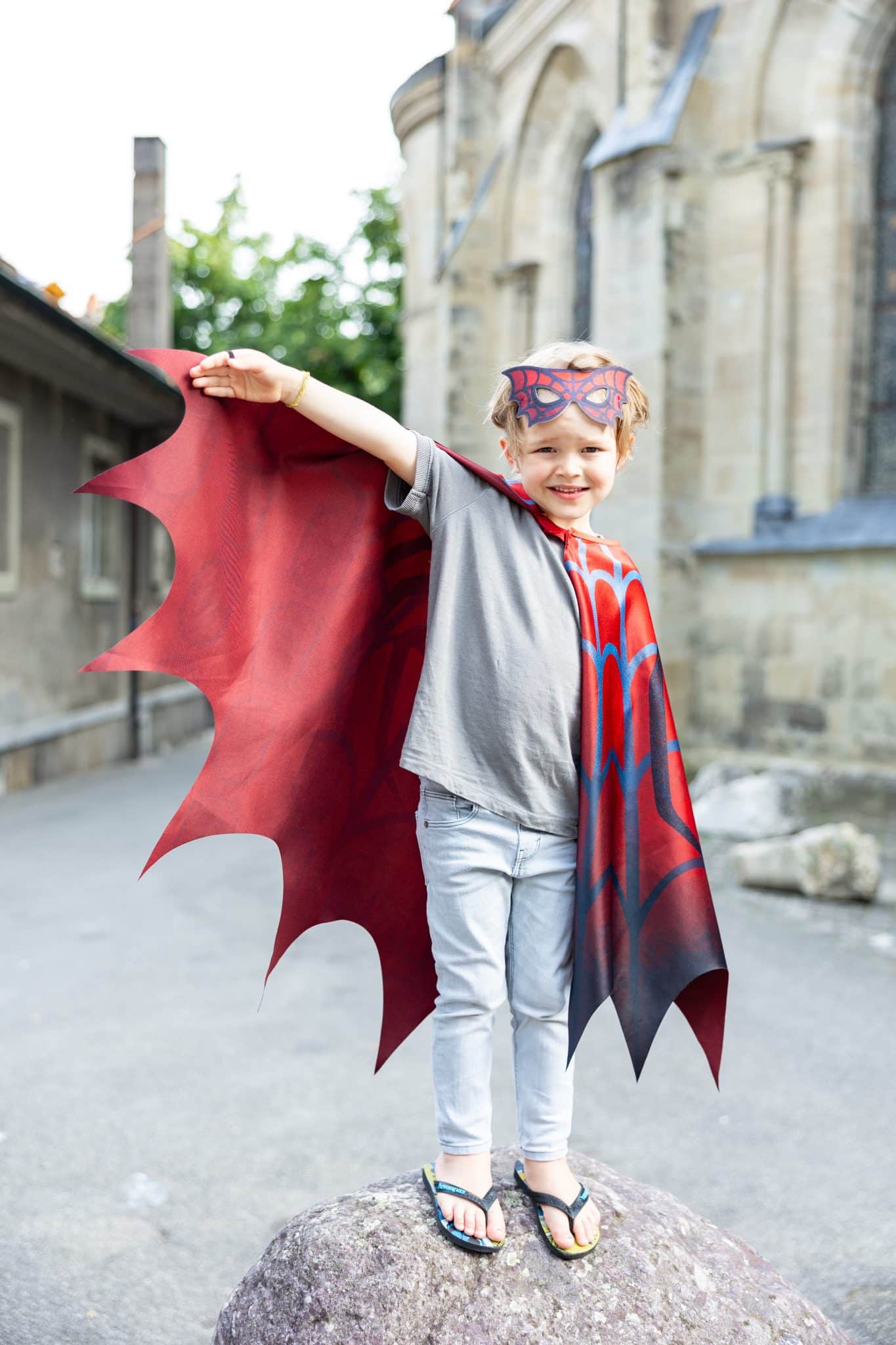 Buiten staat een jongen op een rots met een rode cape spiderman en masker, terwijl hij één arm omhoog houdt.