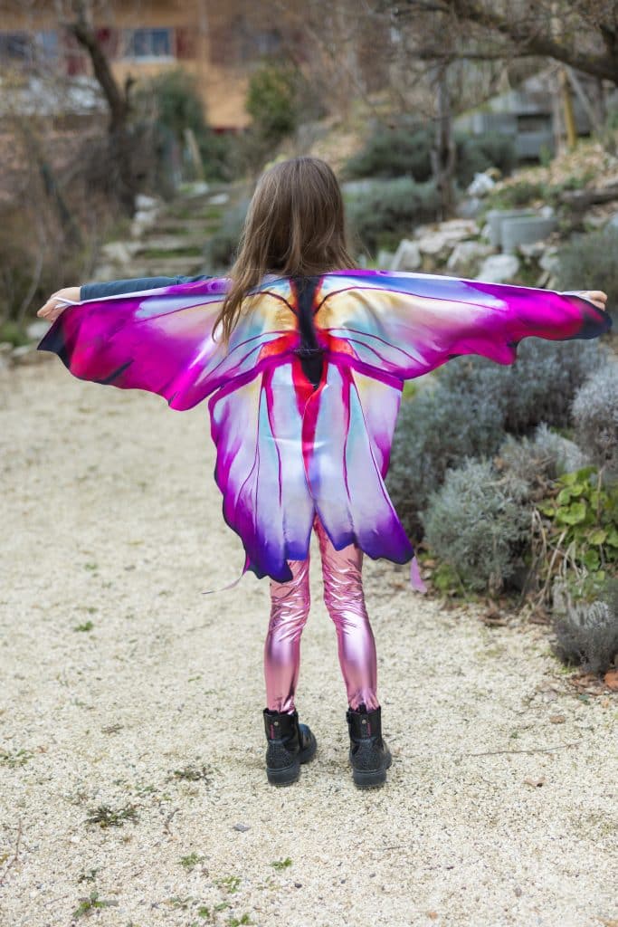 Meisje draagt de paarse vlinder verkleedset met kleurrijke vleugels, van achteren gezien op een tuinpad