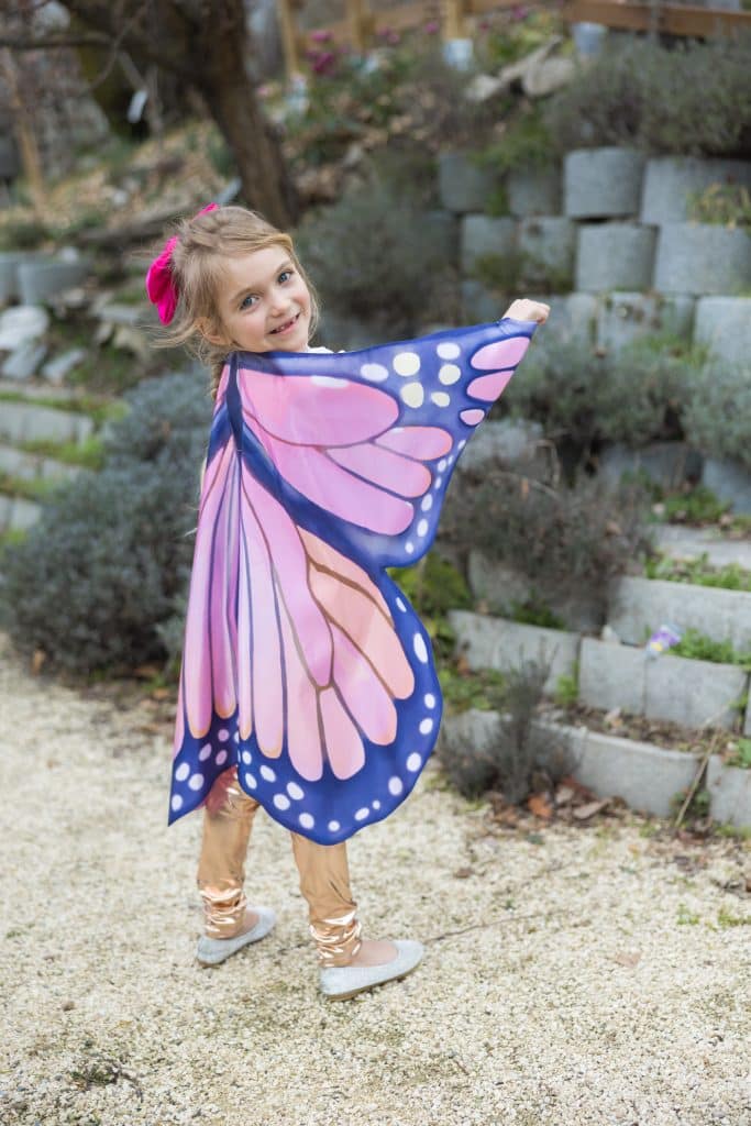 Van voren gezien houdt een meisje één vleugel van de butterfly wings set uitgespreid in de tuin.