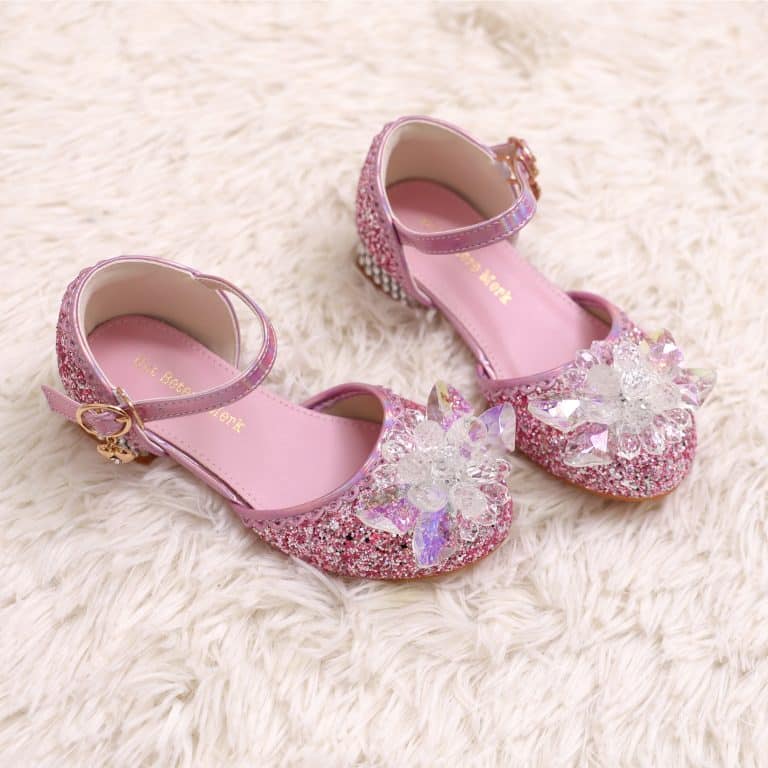 Roze prinsessen schoenen kind met glitters, enkelbandjes en grote bloemversieringen op een zachte witte ondergrond.