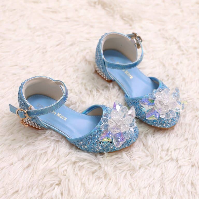 Blauwe prinses schoenen met hakjes en enkelband, bedekt met glitter en grote bloemversieringen op een zachte ondergrond