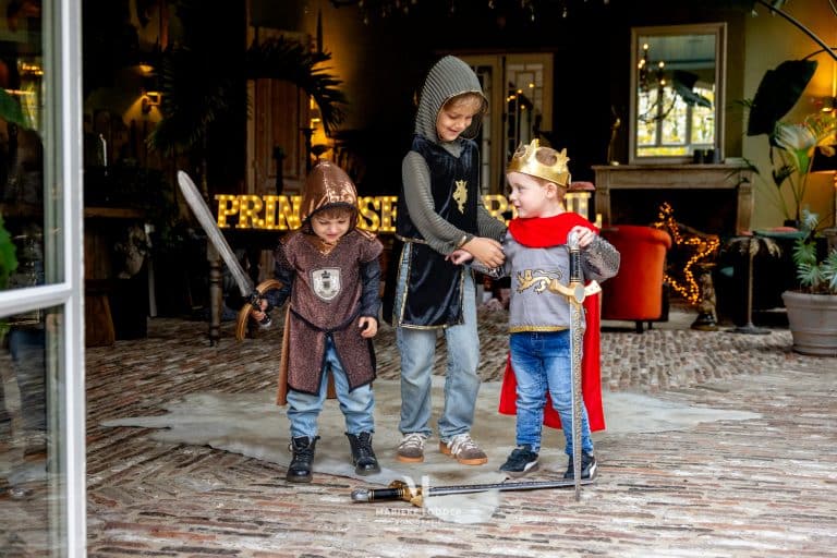 Kind draagt het rode en bruine Souza ridderkostuum met gouden kroon, schildembleem en rode cape, naast twee ridders.