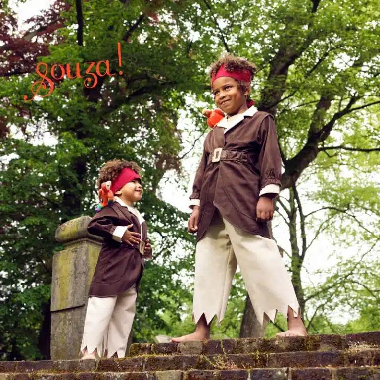 Twee kinderen dragen bruine piraten verkleedkleren met crèmekleurige broek, rode hoofdband en oranje papegaai op de schouder.