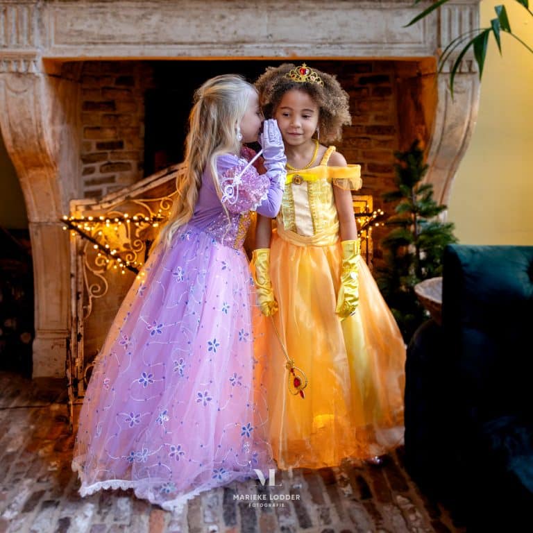 Twee kinderen dragen een paarse Rapunzel prinsessenjurk en gele Belle-jurk met handschoenen en accessoires bij een haardvuur.