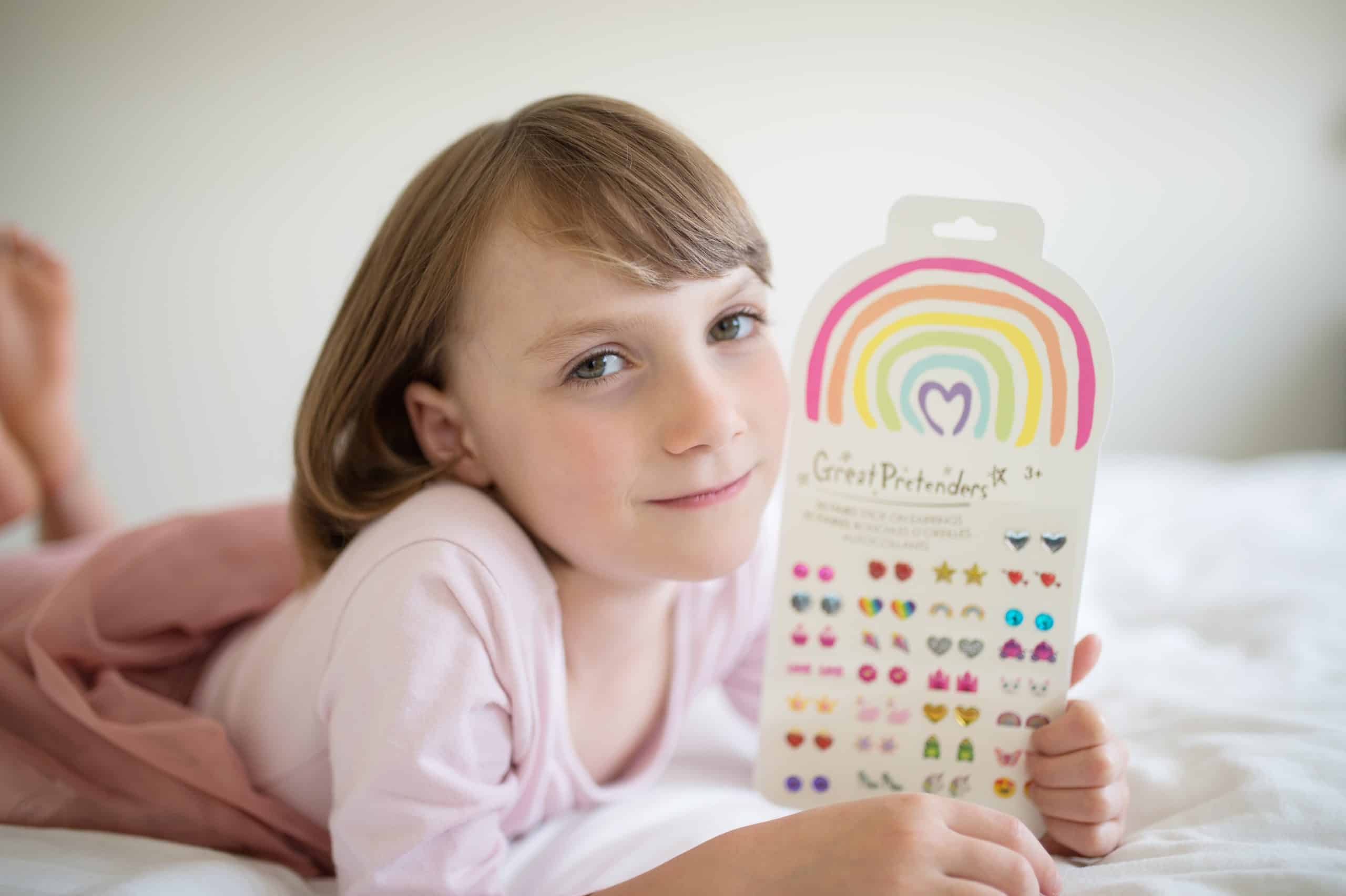 Meisje toont een vel met 30 paar plak oorbellen met regenboog, hartjes en andere kleurrijke figuren