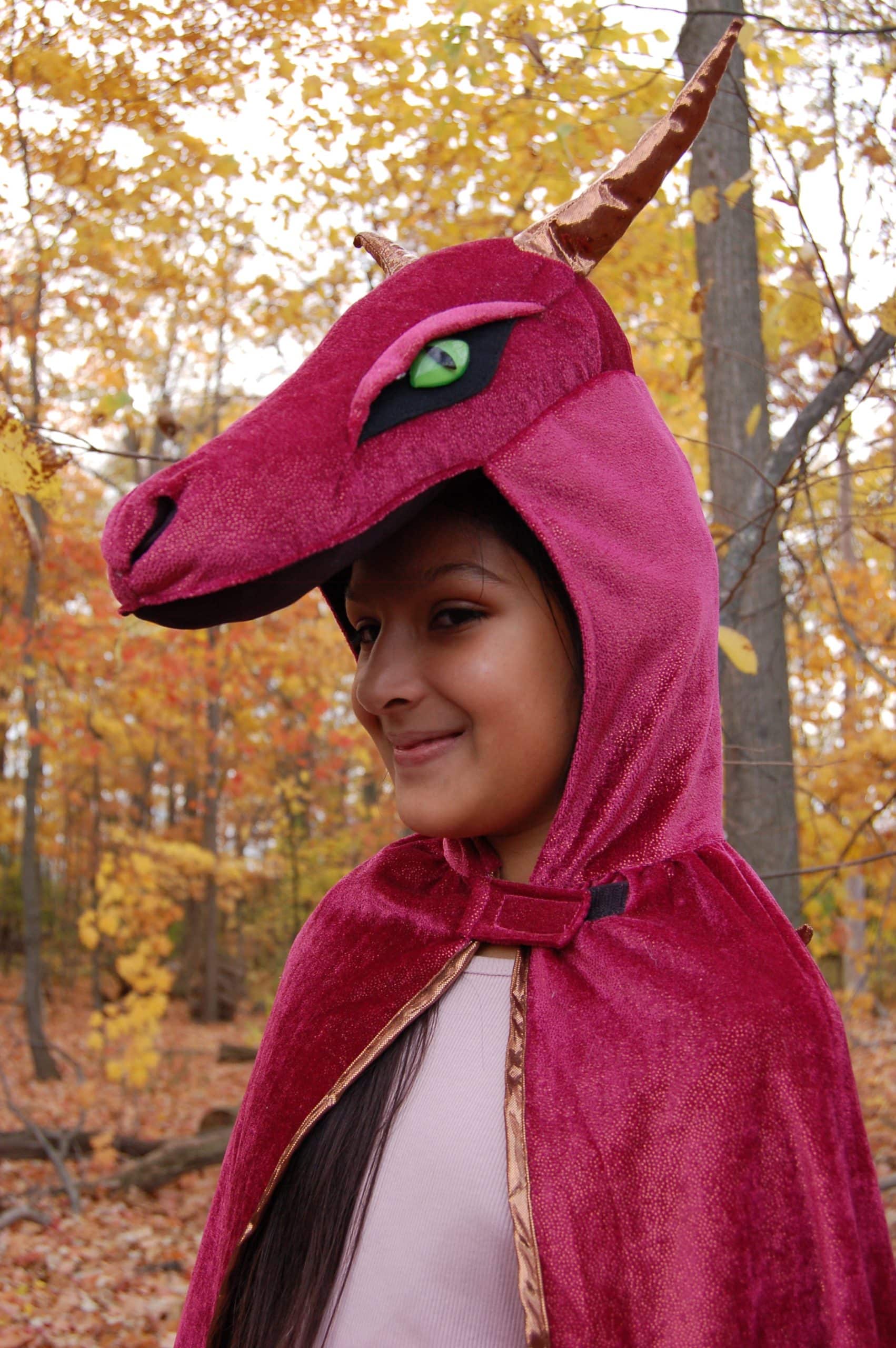 Meisje draagt een rode drakencape met drakenkop, groene ogen en bruine hoorns in een herfstbos.