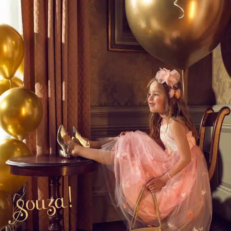 Meisje draagt een elise roze prinsessenjurk met tule rok en roze strik, zittend met haar benen op een tafel.