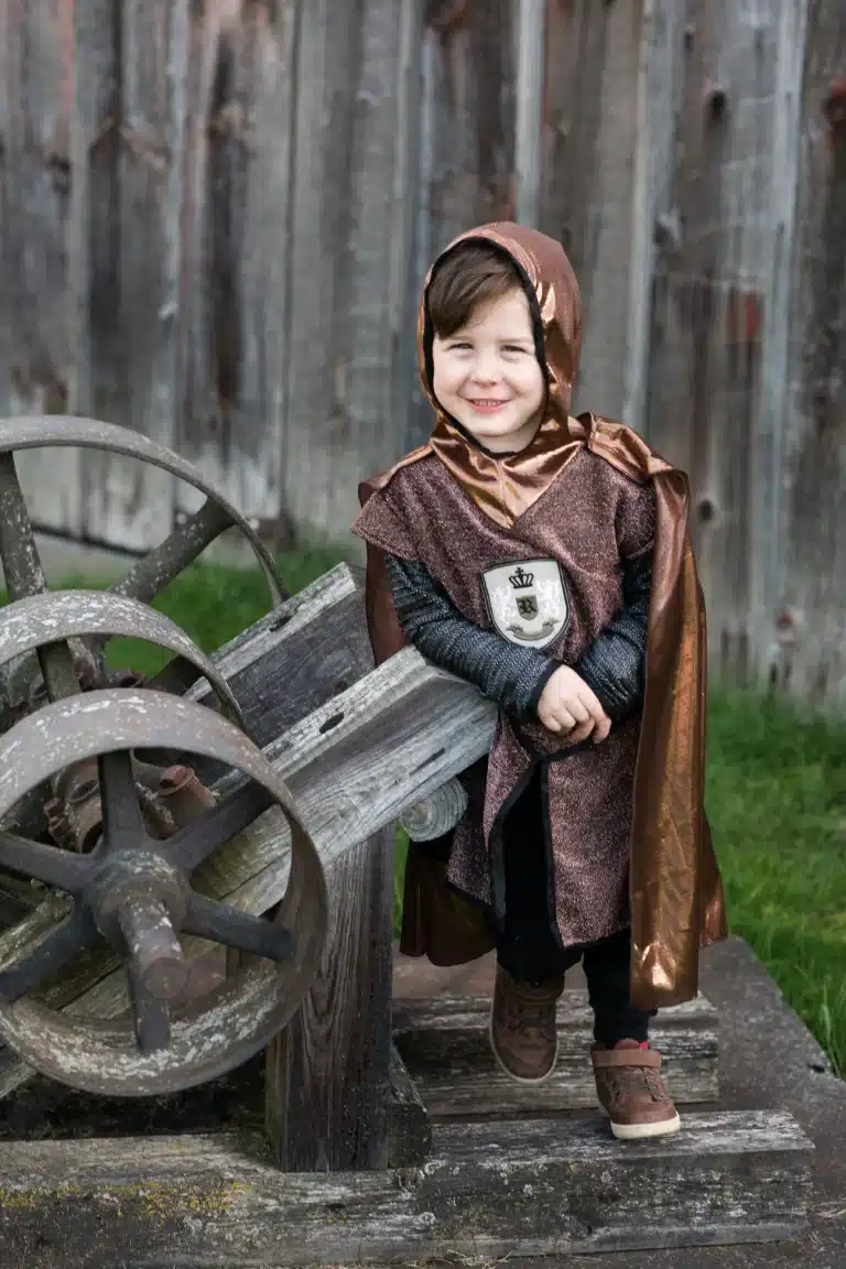 Kind draagt Great Pretenders verkleedkleding ridder met bruine cape, capuchon en ridderembleem naast een houten kanon.