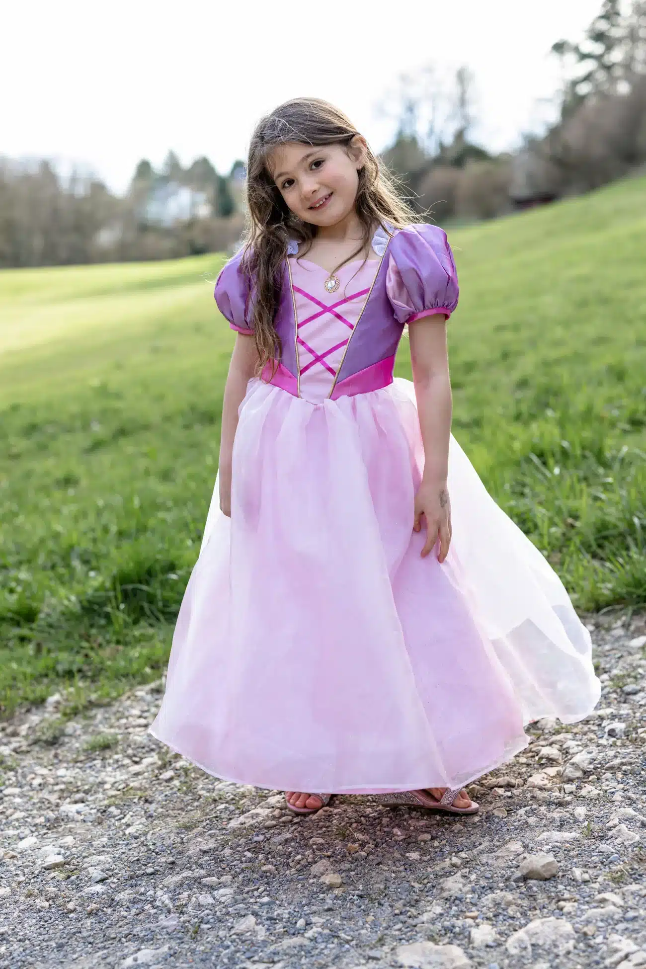 Meisje draagt een paarse Rapunzel prinsessenjurk met roze rok, pofmouwen en gekruiste bandjes, buiten op een grindpad.