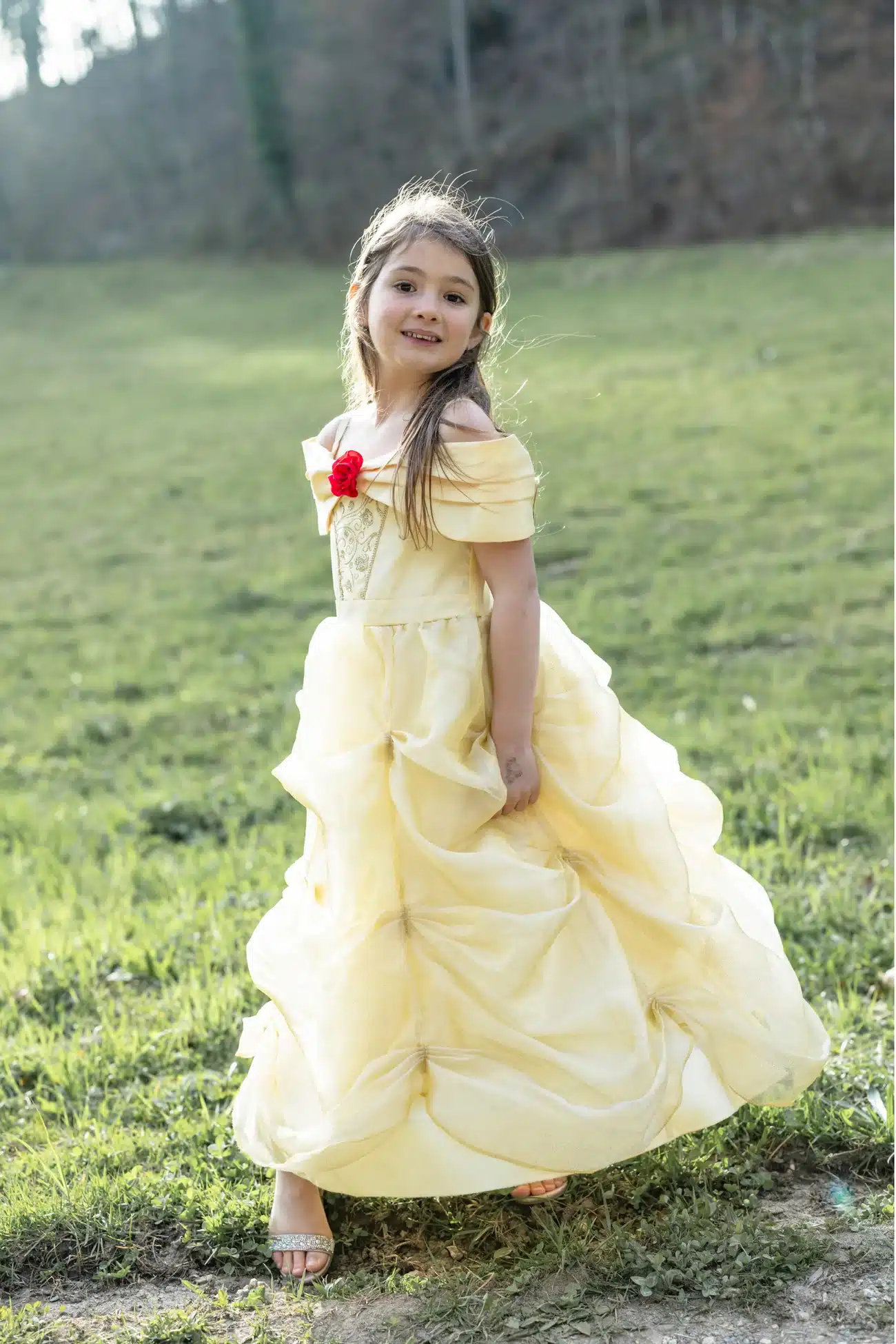 Meisje draagt een gele Belle jurk met gelaagde rok, off-shoulder mouwen en rode roos op een grasveld