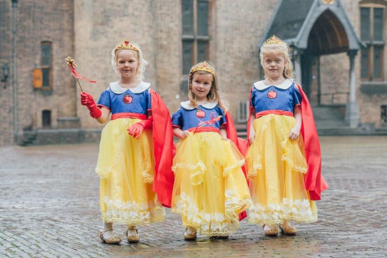 Drie meisjes dragen gele en blauwe Sneeuwwitje jurken met rode capes en kroontjes op een plein.