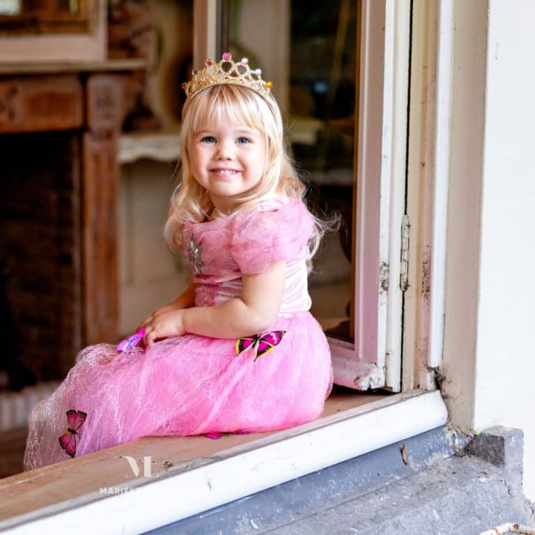 Meisje draagt een roze Assepoester jurk met tiara en vlinderdetails, zittend in een deuropening
