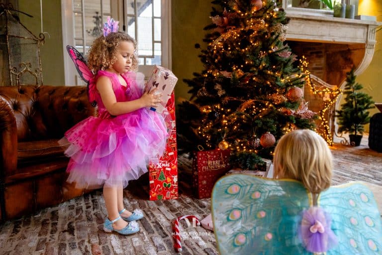 Kind in roze feeënverkleedkleding met vleugels en tule rok bij een versierde kerstboom; op de voorgrond zijn extra vleugels zichtbaar.